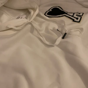 Vit hoodie från Ami Paris - Säljer en stilren vit hoodie från Ami Paris med en unik svart och vit hjärtapplikation på bröstet. Hoodien har en klassisk passform med känguruficka och justerbar huva. Perfekt för en avslappnad look.