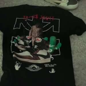Svart Travis Scott x Off-White t-shirt - Säljer en svart t-shirt med Travis Scott-motiv och Off-White-logga. T-shirten har en stor grafisk design på baksidan med gröna och bruna detaljer samt text i rött och vitt. Perfekt för streetwear-entusiaster!