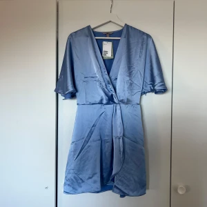 Blå omlottklänning - Snygg blå omlottklänning i satin med korta ärmar. Perfekt för en elegant look med sin v-ringade design och mjuka glans. En stilren och chic klänning för speciella tillfällen.🤍🫧