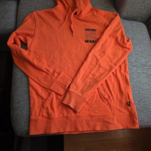 Orange hoodie från Vans - Säljer en cool orange hoodie från Vans med tryck både fram och bak. Hoodien har en klassisk känguruficka och justerbar huva med dragsko. Perfekt för en avslappnad stil. En liten reva finns nere på ena ärmen 