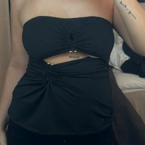 Svart tubtopp med detaljer - Snygg svart tubtopp med rynkade detaljer och små guldfärgade pärlor. Perfekt för en kväll ute eller en festlig tillställning. Toppen har en unik design med en asymmetrisk look.