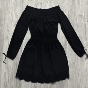 Svart off-shoulder klänning - Snygg svart off-shoulder klänning med långa ärmar och knytband vid ärmsluten. Klänningen har en elastisk midja och spetsdetaljer vid fållen. Underkjol för bekvämlighet.