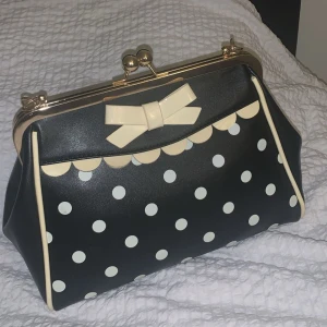 Svart handväska med prickar och rosett - Charmig svart handväska med vita prickar och en söt rosett på framsidan. Väskan har en metallram och en praktisk dragkedja på baksidan. Perfekt för att ge en lekfull touch till din outfit.