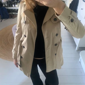 Beige trenchcoat med knappar - Snygg beige trenchcoat med knappar och bälte i midjan. Jackan har långa ärmar och en klassisk krage. Storlek xs från Zara. Finns en lite svart defekt