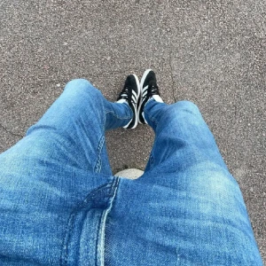 Adidas Gazelle i svart och vit - Snygga Adidas Gazelle sneakers i svart mocka med vita detaljer och den klassiska tre-randiga designen. Skorna har en vit sula och texten 'Gazelle' i guld på sidan. Perfekta för en stilren look.