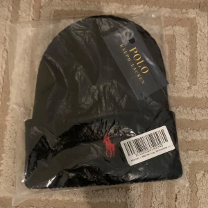 Svart mössa från Polo Ralph Lauren - Snygg svart mössa från Polo Ralph Lauren med det ikoniska röda broderade logotypen framtill. Perfekt för att hålla värmen under kyliga dagar. Det finns 2 st att köpa