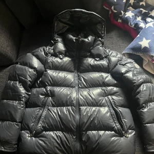 Svart dunjacka från Moncler - Säljer en snygg svart dunjacka från Moncler. Jackan har en dragkedja framtill och en hög krage med huva. Perfekt för kalla dagar med sin pufferdesign som håller värmen. Klassisk och stilren, ett måste i garderoben!