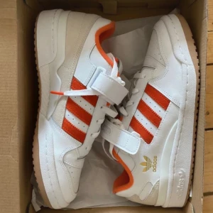 Vita och orange sneakers från Adidas - Snygga vita sneakers med orange detaljer från Adidas. Skorna har klassiska tre ränder i orange och en kardborreband för extra stil och komfort. Perfekta för en sportig look.