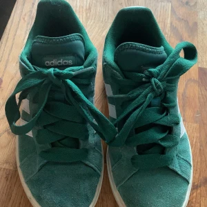 Gröna sneakers från Adidas - Snygga gröna sneakers från Adidas med vita detaljer och snörning. Perfekta för en avslappnad stil. Skorna har en klassisk design med tre vita ränder på sidan och Adidas-logga på hälen.