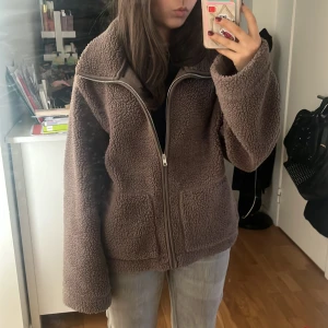 Brun teddyjacka med dragkedja - Mysig brun teddyjacka med dragkedja framtill. Jackan har en hög krage och långa ärmar, perfekt för kyliga dagar. Den har en avslappnad passform och är superbekväm att bära.