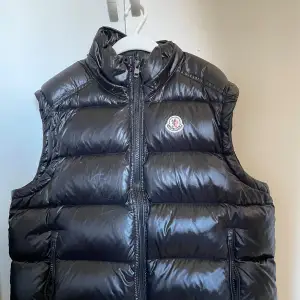 Snygg svart dunväst från Moncler med dragkedja framtill. Västen har en hög krage och är perfekt för kyligare dagar. Den har en stilren design med Moncler-logga på bröstet.