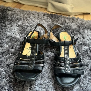 Svarta sandaler med remmar - Snygga svarta sandaler från Rieker med flera remmar och dekorativa detaljer på framsidan. Sandalerna har en bekväm sula med mönstrad undersida för bra grepp. Perfekta för varma sommardagar.