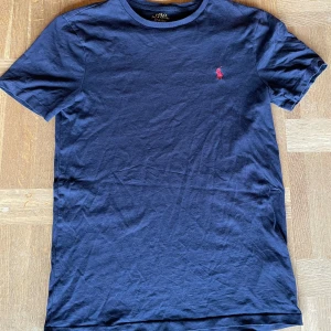 Mörkblå t-shirt från Ralph Lauren - Säljer en stilren mörkblå t-shirt från Ralph Lauren med en broderad röd logotyp på bröstet. Säljer för att den är för liten för mig. Tröjan är i bra skick och har endast ett litet hål vid kragen men ingenting man tänker på. Den är i stl S och slim fit. Kom gärna med frågor.