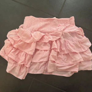 Rosa volangkjol i XS - Supersöt kort rosa kjol med flera lager volanger och smock i midjan. Kjolen är lätt och luftig, perfekt för varma dagar. Passar dig som gillar en feminin och lekfull stil.