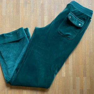 Mörkgröna velourbyxor Juicy Couture - Mörkgröna mjukisbyxor från Juicy Couture i mjuk velour. Byxorna har raka ben, fickor med lock och silverfärgade knappar samt broderad JC-logga på ena fickan. Dem är uppsydda en liten bit med lätta att sprätta upp.