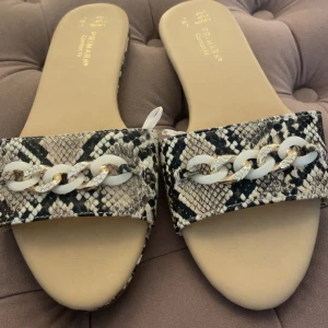 Ormskinnsmönstrade sandaler Primark - Snygga sandaler från Primark med ormskinnsmönster i beige och svart. Dekorerade med en chunky kedja i vitt och guld med glittriga detaljer. Platt sula och öppen tå. 