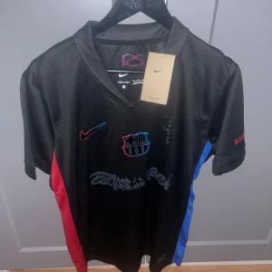 FC Barcelona x Nike svart fotbollströja - Snygg svart FC Barcelona x Nike fotbollströja med korta ärmar. Tröjan har röda och blå sidopaneler, klubbmärke på bröstet och Nike-logga i rött. Materialet är lätt och andas, perfekt för match eller träning. Unik Cactus Jack-grafik framtill by Travis Scott. True to size. Pris kan diskuteras mer privat 👀