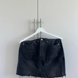 Svart jeanskjol med fransig kant - Snygg svart jeanskjol med klassiska fickor fram och bak. Kjolen har rå, fransig nederkant för en cool vibe och stängs med knapp och dragkedja framtill. Perfekt att styla med sneakers eller boots för en avslappnad look.