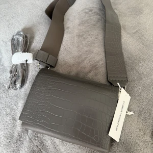 Ny liten axelväska med lock - •Väskan har bred, justerbar axelrem med stort, silverfärgat spänne. •••Remmen är avtagbar och kan bytas ut mot smal rem som medföljer. •Under locket finns ett huvudfack med dragkedjeficka och kortfack inuti. •••Ordinarie pris: 499 kr  • Liten väska i blank polyester • Locket låses med magnetknapp • 1 huvudfack, 1 innerfack med dragkedja, 1 kortficka • 2 axelremmar  Mått:  Bredd: 18,5 cm Höjd: 13 cm  •••Djurfritt och rökfritt hem