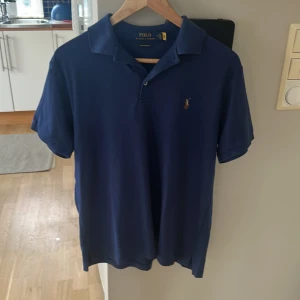 Blå pikétröja från Polo Ralph Lauren - Snygg marinblå pikétröja från Polo Ralph Lauren i bomull. Klassisk krage, korta ärmar och broderad logga på bröstet. Modellen är custom slim fit och passar perfekt för en stilren look.