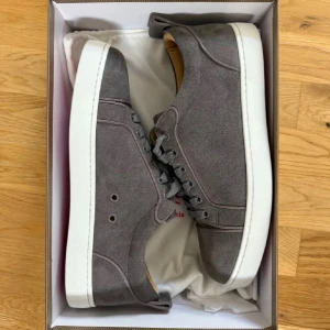 Grå mockasneakers från Christian Louboutin - Snygga grå sneakers i mocka från Christian Louboutin med klassisk vit sula och ikonisk röd undersida. Skorna har rund tå, snörning och diskret branding på insidan. Perfekta för dig som vill ha en stilren och lyxig look.