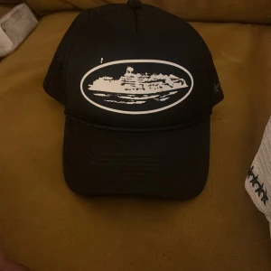 Svart truckerkeps från Corteiz - Svart truckerkeps från Corteiz med vit broderad grafik framtill och mesh på sidorna och bak. Kepsen har broderade detaljer med taggtrådsmotiv och logga på insidan. Justerbar passform och klassisk böjd skärm.