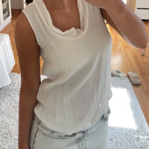 Vit blus från Zara  - Säljer en vit ärmlös blus från Zara med snygg draperad krage och knytning i nacken. Baksidan har dekorativa knappar i olika färger som ger en unik touch. Tunt, lätt material som känns luftigt och fräscht. Perfekt till jeans eller kjol!