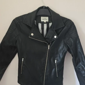 Svart skinnjacka från KappAhl, 164 - Snygg svart skinnjacka från KappAhl i storlek 164. Jackan har klassisk bikerstil med dragkedja framtill, två fickor med dragkedjor och coola detaljer på axlarna. Perfekt för dig som vill ha en edgy look till vardagsoutfiten.