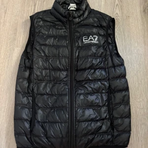 Svart dunväst från Emporio Armani EA7 - Snygg svart dunväst från Emporio Armani EA7 med quiltad design och hög krage. Västen har dragkedja framtill och logga broderad på bröstet. Perfekt att slänga över en hoodie för en sportig och clean look.