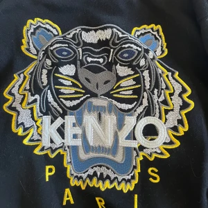 Svart Kenzo sweatshirt - Svart kenzo sweatshirt  med ikonisk broderad tiger i grått, blått och gult framtill. Tröjan har rund halsringning och ribbade muddar. Perfekt statement-plagg för dig som vill sticka ut med cool design och snygga detaljer. (Inte äkta!)