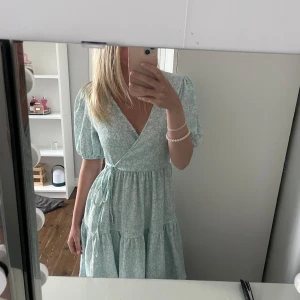 Ljusblå omlottklänning med puffärm - Superfin ljusblå omlottklänning med småblommigt mönster och puffiga kortärmar. Klänningen har knyt i midjan och volangdetaljer i kjolen, perfekt för sommarens alla tillfällen. Sval och luftig känsla.