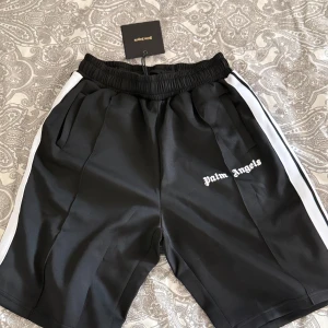 Palm Angels svarta shorts - Svarta shorts från Palm Angels med vita sidoränder och broderad logga på vänster ben. Elastisk midja med snörning och sportig vibe. Perfekta för chill dagar eller streetwear-stil. Tillverkade i ett mjukt och följsamt polyestermaterial.