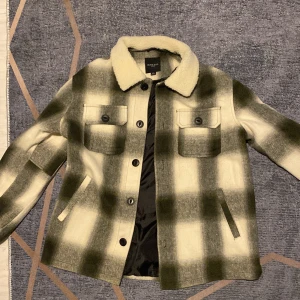 Rutig overshirt från Brave Soul med teddykrage - Snygg rutig overshirt från Brave Soul i beige och mörkgrön med mjuk teddykrage. Jackan har två bröstfickor med knappar, sidofickor och helknäppning framtill. Fodrad insida för extra komfort. Perfekt för lager-på-lager-stil.