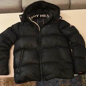 Svart Tjock dunjacka Tommy Hilfiger - Svart pufferjacka från Tommy Hilfiger med huva och dragkedja. Jackan har två snedställda fickor med dragkedja och diskret logga på bröstet samt ärmen. Insidan av kragen har tydlig Tommy Hilfiger-text. Perfekt för kalla dagar. Några tecken på användning.