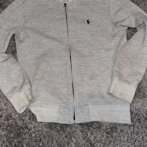 Grå ziptröja från Polo Ralph Lauren XS - Snygg grå ziptröja från Polo Ralph Lauren i mjuk bomullsblandning. Klassisk design med svart broderad logga på bröstet, ribbade muddar och två fickor framtill. Perfekt för lager-på-lager och chill vardagsstil.