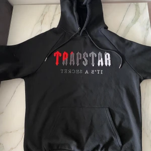 Svart Trapstar hoodie med brodyr - Svart hoodie från Trapstar med broderad logga i rött, grått och vitt på bröstet. Klassisk känguruficka framtill, huva med snörning och mjukt material på insidan. Perfekt för dig som gillar streetwear och vill sticka ut med en fet logga.