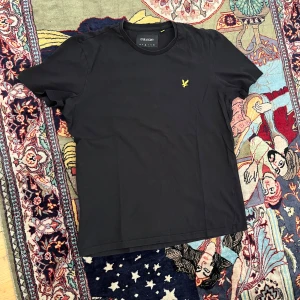 Svart t-shirt från Lyle & Scott - Svart t-shirt från Lyle & Scott med klassisk rund hals och korta ärmar. På bröstet sitter den ikoniska gula örnloggan broderad. T-shirten är i mjuk bomull och har en clean, stilren look som funkar till allt.