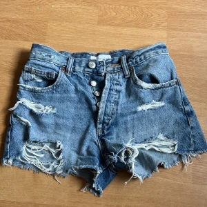 Blå slitna jeansshorts från Zara - Riktigt snygga blå jeansshorts från Zara med hög midja, slitna detaljer och fransig kant. Klassisk femficksmodell med knappar framtill och coolt ruffig look. Perfekta för varma dagar när du vill ha en avslappnad vibe.