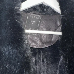 Svart pälsjacka från Guess - Snygg svart pälsjacka från Guess med bred fuskpäls-krage och coolt mönstrad yta. Jackan har dragkedjor på fickorna och insidan är fodrad med mjuk pälsimitation. Perfekt för dig som vill sticka ut med en statement-jacka i vinter.