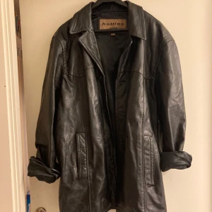 Svart skinnjacka från Hollies - Säljer en svart skinnjacka från Hollies i oversized modell. Jackan har klassisk krage, knäppning framtill med knappar och två stora fickor. Saknar dock två knappar framtill, se bild 4. Hål i en ficka. Perfekt för dig som gillar en edgy och tidlös look. 