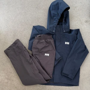 Mörkblå regnjacka från Helly Hansen - Mörkblå regnjacka från Helly Hansen med huva och tryckknappar framtill. Jackan har en enkel, sportig design med HH-logga på bröstet. Perfekt för regniga dagar och outdoor-aktiviteter.