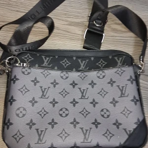 axelväska  - Snygg axelväska  i svart och grått med klassiskt monogrammönster. Väskan har justerbar axelrem, tvådelad design med dragkedja och är tillverkad i skinn. Perfekt för dig som vill ha en stilren och lyxig accessoar