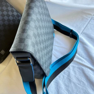 Louis Vuitton rutig axelväska med blått foder - Snygg axelväska från Louis Vuitton i klassiskt grått och svart rutigt mönster. Väskan har en bred justerbar axelrem i blått och svart, samt ett blått innerfoder. Perfekt för dig som vill ha en stilren och praktisk väska med lyxig känsla.