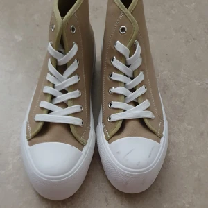 Sneakers - Stilrena höga sneakers i beige canvas med vita gummisulor och tåhätta. Skorna har vita snören och silverfärgade öljetter, samt diskreta kontrastsömmar. Perfekta för dig som gillar en clean och klassisk look med modern touch.
