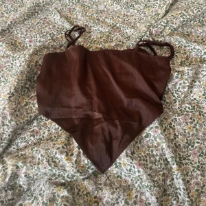 Brun scarf-topp från H&M Divided XS - Säljer en brun scarf-topp från H&M Divided i storlek XS. Toppen har smala axelband, glansig satinlook och smockad rygg för extra komfort. Snygg asymmetrisk form med spetsig nederkant, perfekt för att lyfta din outfit.