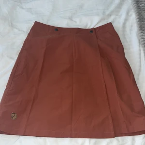 Rostbrun omlottkjol från Fjällräven - Rostbrun omlottkjol från Fjällräven med inbyggda shorts och klassisk logga nertill. Kjolen har två knappar i midjan och är tillverkad i slitstarkt material, perfekt för aktiva dagar. Snygg och praktisk design med sportig känsla.