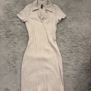 Beige ribbad kortklänning med krage - Supersnygg beige kortklänning från Bershka med ribbad struktur, v-ringning och klassisk krage. Klänningen har korta ärmar och figurnära passform som framhäver siluetten. Perfekt för dig som gillar stilrena och trendiga plagg.