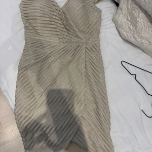 Glittrig champagnefärgad kortklänning - Supersnygg kortklänning i champagnefärg med glittrigt tyg och omlott-design. Klänningen har en axelbandslös look och diagonala ränder. Färgen lutar lite åt det guldiga hållet. Är i storlek 38 men funkar även som en 36a. Pris går att diskuteras!