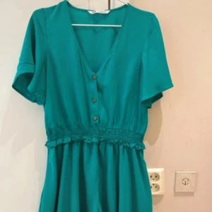  Ny Turkos kortklänning med knappar - Snygg turkos kortklänning med v-ringning, knappar framtill och smock i midjan. Klänningen har korta ärmar och ett flowy fall som ger en avslappnad men trendig look. Perfekt för varma dagar och enkel att styla med sneakers eller sandaler.