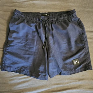 Marinblå badshorts från J.Lindeberg - Snygga marinblå badshorts från J.Lindeberg med elastisk midja och snörning. Klassisk design med sidofickor och en bakficka. Materialet känns lätt och passar perfekt för varma dagar. J.Lindeberg-logga framtill ger en clean och sportig vibe.